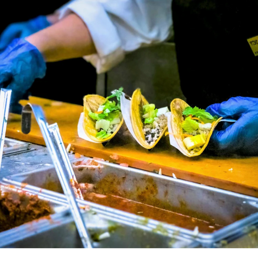 Food service worker wearing blue nitrile gloves preparing tacos at a restaurant counter with fresh ingredients.