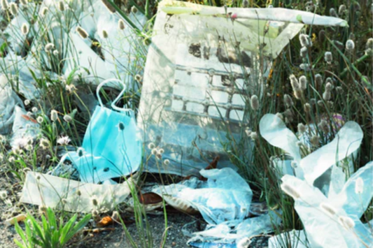Plastic waste, including disposable bags and a face mask, scattered among grass and wild plants, highlighting environmental pollution and littering issues.